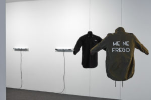Deborah Rundle, Political Colours, 2018. Black shirt with printed text, military patch and officers’ stars, khaki jacket with printed text, mannequin torsi. Commissioned by Te Tuhi, Auckland. Optimism of the Will, 2018. Two fluorescent tubes and batten holders, vinyl cut text. Photo by Sam Hartnett.